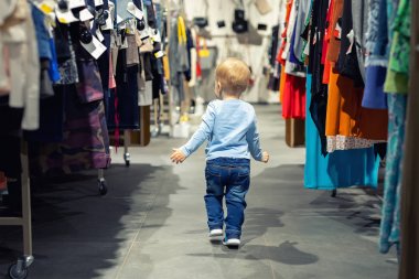 Sevimli kafkas sarışın bebek çocuk askıları ile raf arasında giysi perakende mağazasında tek başına yürüme. Bebek yetişkin alışveriş dünyasını keşfeder. Bebek büyük hipermarket alışveriş merkezinde kaybolmak