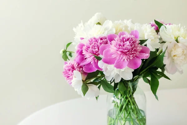 Close-up bouquet of fresh big pink, white and cream peonies in simple glass jar on glance table indoor. Vase with beautiful tender spring flowers on glass table — Stock Photo, Image