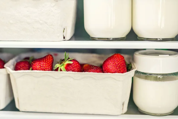 Refrigerator shelf with organic juicy strawberries in bio recycled paper boxes and homemade natural milk yogurt. Fridge filled with berries in cardboard containers. Healthy and eco friendly food diet