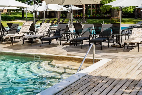 Junto a la piscina con agua azul clara y tumbonas bajo sombrillas en el calor del día de verano. Piscina en el complejo en el verde bosque de pinos de coníferas. Lugar de destino fin de semana — Foto de Stock