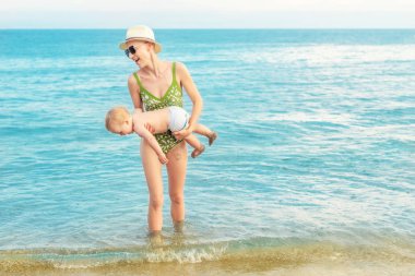 Sıcak yaz yağmuru ile tropikal plajda açık mavi su dışarı sevimli Beyaz Toddler oğlan taşıyan genç güzel kel anne. Anne deniz veya okyanus bebek oğul ile eğlenmek. Seyahat tatil ve tatil