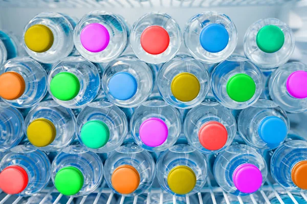 Rows of many transparent plastic drinking water bottles with bright multiclored colorful lids in white refrigerator. Mineral water stack storage in fridge. Healthcare and dehydration prevention