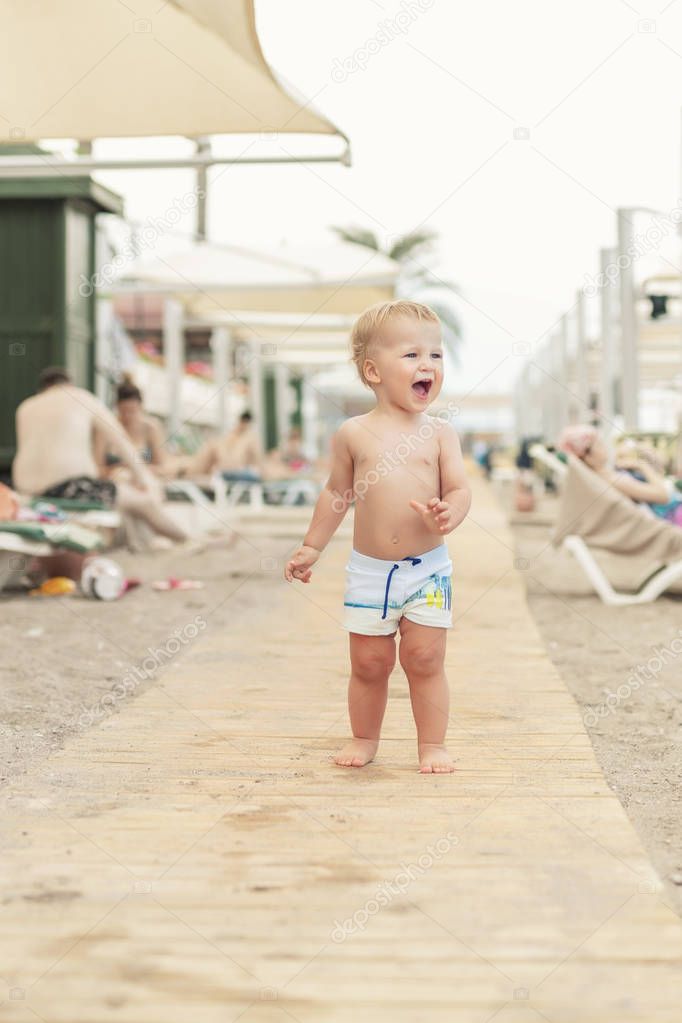 Cute caucasian toodler boy walking alone on sandy beach between chaise-lounge. Adorable happy child having fun playing at seaside shore during vacation trip