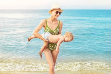 Sıcak yaz yağmuru ile tropikal plajda açık mavi su dışarı sevimli Beyaz Toddler oğlan taşıyan genç güzel kel anne. Anne deniz veya okyanus bebek oğul ile eğlenmek. Seyahat tatil ve tatil