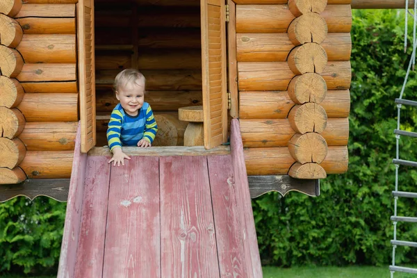 Lindo adorable niño caucásico que se divierte deslizándose por el tobogán de madera en el patio natural ecológico en el patio trasero en otoño. Retrato infantil divertido juguetón disfrutando de actividades deportivas al aire libre —  Fotos de Stock