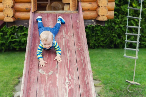 Lindo adorable niño caucásico que se divierte deslizándose por el tobogán de madera en el patio natural ecológico en el patio trasero en otoño. Retrato infantil divertido juguetón disfrutando de actividades deportivas al aire libre —  Fotos de Stock