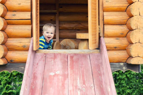 Lindo adorable niño caucásico que se divierte deslizándose por el tobogán de madera en el patio natural ecológico en el patio trasero en otoño. Retrato infantil divertido juguetón disfrutando de actividades deportivas al aire libre —  Fotos de Stock