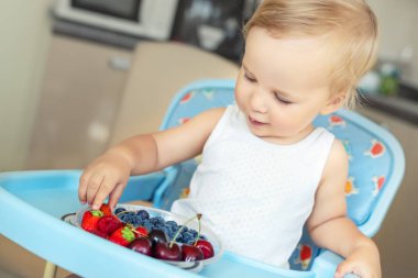 Sevimli sevimli kafkas sarışın çocuk ev mutfağında mama sandalyesinde oturan farklı mevsimsel taze olgun organik çilek tatma keyfini çıkarın. Mutlu çocuk arkadaşı ile doğal tatlı sağlıklı gıda paylaşımı