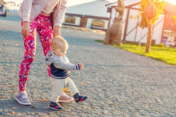 Cuki kis aranyos szőke kisgyermek fiú hogy első lépések az anya támogatást a városi parkban este naplemente idő. Boldog vicces gyermek tanulás járni anyu segítségével kültéri — Stock Fotó