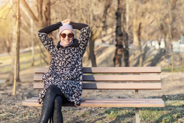 Mulher branca feliz bonita na jaqueta, chapéu e óculos de sol desfrutar sentado no banco no parque da cidade ou na floresta no dia ensolarado de outono. Sorrindo menina adulta jovem se divertindo durante a temporada de outono andando — Fotografia de Stock