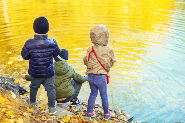 Tres niños amigos jugando a la pesca con palos cerca del estanque en el hermoso parque de otoño de oro. Hermanitos divirtiéndose cerca del lago o el río en otoño. Feliz infancia y niño concepto de recreación al aire libre — Foto de Stock