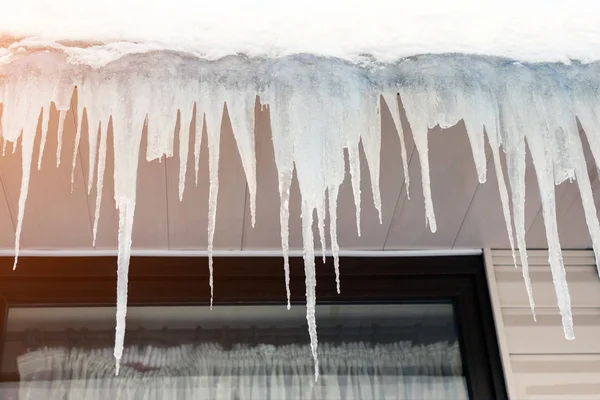Nagy átlátszó kék jégcsapok akasztott a tetőn épület ablak háttérben. Téli vagy tavaszi olvadáspont szezonális veszély — Stock Fotó