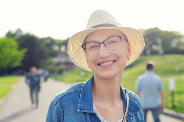 Feliz joven mujer calva caucásica con sombrero y ropa casual disfrutando de la vida después de sobrevivir al cáncer de mama. Retrato de la hermosa chica sin pelo sonriendo durante el paseo en el parque de la ciudad después de curar la enfermedad —  Fotos de Stock