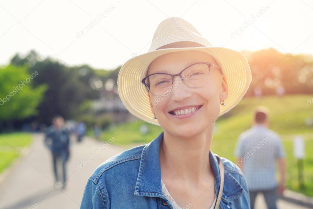 Happy young caucasian bald woman in hat and casual clothes enjoying life after surviving breast cancer. Portrait of beautiful hairless girl smiling during walk at city park after curing disease