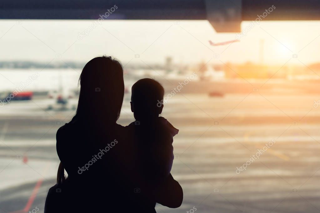 Silhouette of mother holding on hands little toddler boy with window of airport on background. Departure and arrival. Single mother with child emigration. Mom with son meeting or seeing off father