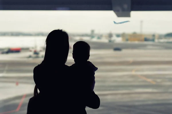 Silueta de la madre sosteniendo en las manos niño pequeño con ventana del aeropuerto en el fondo. Salida y llegada. Madre soltera con emigración infantil. Mamá con hijo reunión o despedida padre —  Fotos de Stock