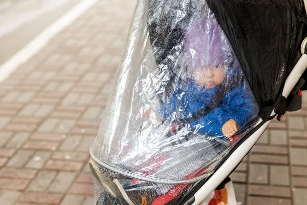 Kleine schattige Kaukasische peuter jongen in casual sport kleding hebben plezier genieten van wandelen tijdens de regen in de herfst dag. Kind zittend in kinderwagen met regenbescherming — Stockfoto