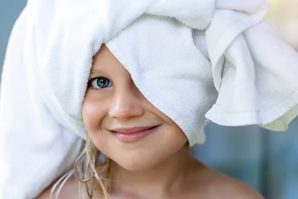 Cute adorable caucasian little blond girl wearing white towel on we head after shower or bathing at bathroom. Portrait of cheerful smiling female child — Stockfoto