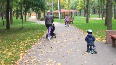 Küçük çocuk asfalt yolda scooter dengeli bisiklete biniyor. Annesiyle birlikte sonbaharda şehir parkında yürüyor. Alışveriş merkezi çocuğu şehir caddesinde hızla bisiklet sürerken eğleniyor.