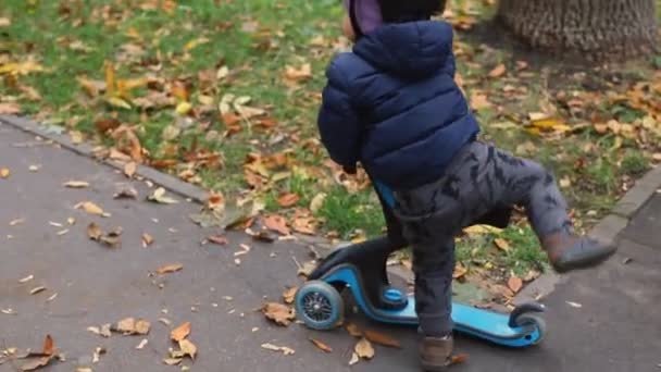 Küçük çocuk asfalt yolda scooter dengeli bisiklete biniyor. Annesiyle birlikte sonbaharda şehir parkında yürüyor. Alışveriş merkezi çocuğu şehir caddesinde hızla bisiklet sürerken eğleniyor. — Stok video