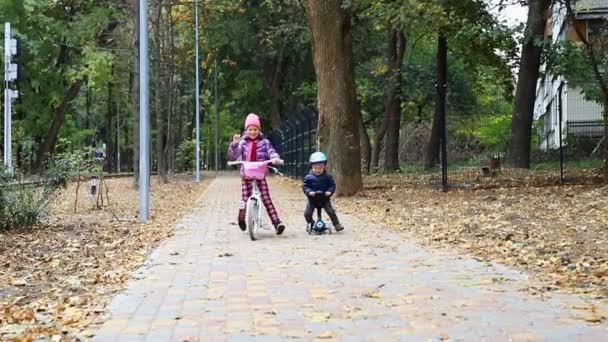 Маленький хлопчик їде на скутері баланс велосипед на асфальтній прогулянці разом з матір'ю, що йде поблизу шляху до міського парку восени на відкритому повітрі. Маленька дитина весело катається на велосипеді швидко на міській вулиці — стокове відео