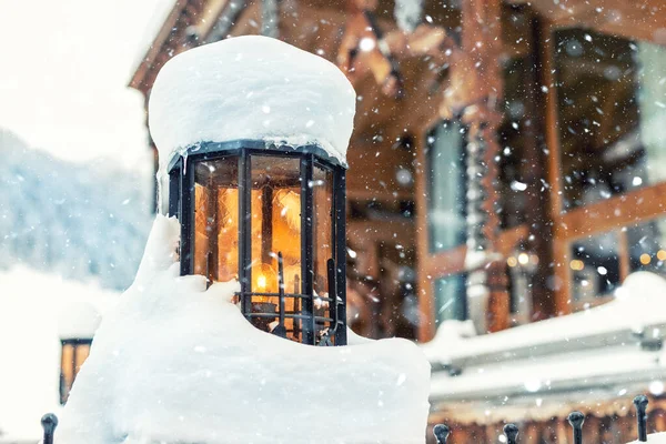 Hangulatos vintage utcai lámpa narancssárga üveggel borított hó és fából készült rusztikus ház a háttérben a hóesés. Retro fém lámpás dekoráció az alpesi resort utcában. Karácsonyi hangulat — Stock Fotó