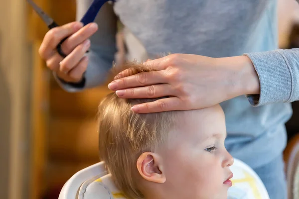 Close-up jonge volwassen blanke moeder maken kapsel fot schattige bewonderenswaardige zoon peuter jongen thuis als gevolg van quarantaine en lockdown. Moeder knippen haar van kind met schaar en haarborstel — Stockfoto