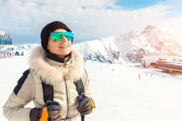 Portrait of beautiful young adult caucasian woman in sport suit, hat, sunglasses and backpack looking and smiling on mountain peak covered with snow on bright sunny winter day at alpine ski resort — Stock Photo, Image