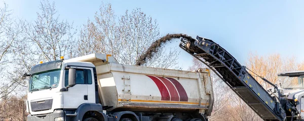 Asphalt milling and grinding machine at road repair and construction site throwing shredded old bitumen in big industrial heavy dumper tipper truck for recycling. Highway renewal with heavy machinery