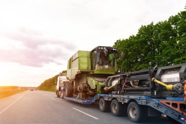 Yarı karavan platformlu ağır sanayi kamyonu gün batımında ya da gün doğumunda ortak otoyolda parçalanmış hasat makinesini birleştirdi. Tarım ekipmanları taşıma hizmeti