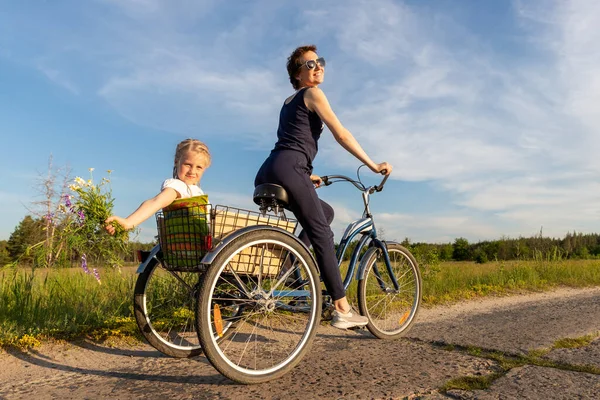 Молодая взрослая кавказская мама наслаждается отдыхом, катаясь на велосипеде с милой очаровательной блондинкой, держащей полевой цветок на живописной сельской дороге в яркий солнечный день. Отпуск за городом — стоковое фото