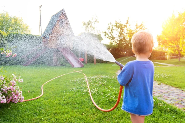 Nette entzückende kaukasische blonde Kleinkind Junge genießen Spaß beim Gießen Garten Blumen und Rasen mit Schlauchleitungen Sprinkler zu Hause Hinterhof an sonnigen Tagen. Kleine Helfer lernen Gartenarbeit im Sommer im Freien — Stockfoto