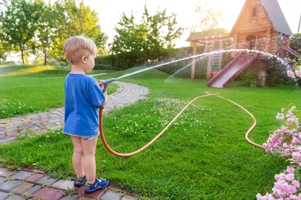 Bonito adorável caucasiano loiro criança menino desfrutar de ter diversão rega jardim flor e gramado com mangueira aspersor em casa quintal no dia ensolarado. Criança pequeno ajudante aprender jardinagem no verão ao ar livre — Fotografia de Stock