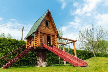Parkın ya da evin bahçesinde merdivenleri ve tahta kaydırağıyla küçük ahşap bir oyun evi kulübesi. Yeşil çimen bahçesi ve açık mavi gökyüzü açık arka planda güneşli yaz gününde.