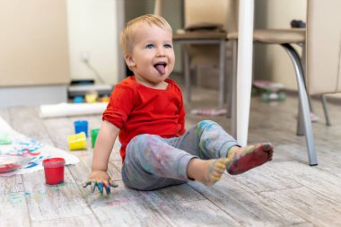 Sevimli, beyaz, sarışın, yaramaz çocuk evde fırça ve avuç içiyle resim yaparken eğleniyor. Mutlu, neşeli çocuk sanat eseri çizerek gülümsüyor. Kirli oda.