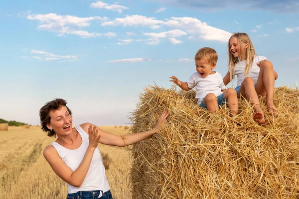 Jovem adulto atraente mãe bonita com filho pequeno e filha gostam de se divertir brincando sentado perto de fardo de feno dourado no campo colhido de trigo perto da fazenda. Crianças felizes na paisagem rural — Fotografia de Stock
