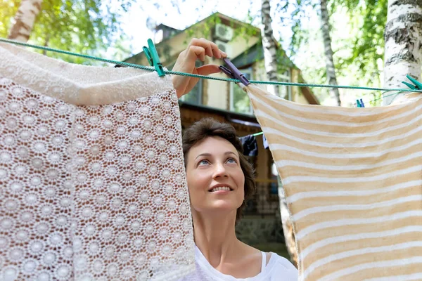 Candid real life portrait of young adult beautiful attractive caucasian woman hanging up fresh washed family clothes on birch tree clothesline with pins at home yard on bright sunny day outdoors — Stock Photo, Image