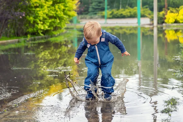 Pequeño niño pequeño rubio caucásico juguetón disfrutar de divertirse jugando saltar en charco sucio con pantalones azules impermeables y botas de lluvia de goma en casa patio calle al aire libre. Concepto de infancia feliz — Foto de Stock