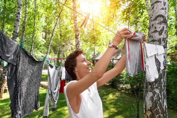 Candid real life portrait of young adult beautiful attractive caucasian woman hanging up fresh washed family clothes on birch tree clothesline with pins at home yard on bright sunny day outdoors — Stock Photo, Image