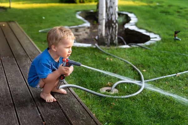 Lindo adorable niño rubio caucásico disfrutar de divertirse regando flor de jardín y césped con aspersor de manguera en el patio trasero del hogar en el día soleado. Niño pequeño ayudante aprender jardinería en verano al aire libre — Foto de Stock