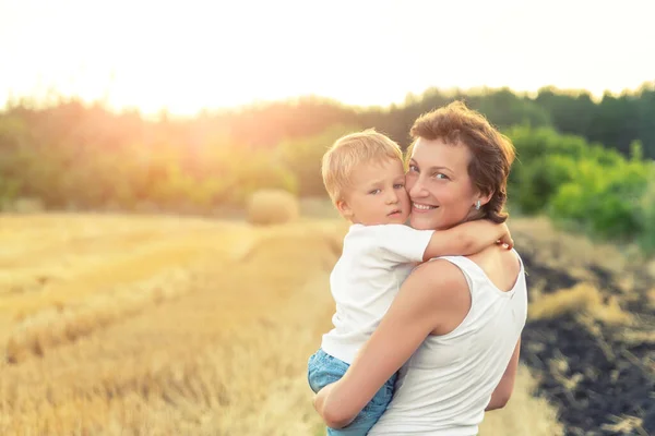 Young adult attractive beautiful mom holding on hand and hugging little son enjoy walking by golden wheat harvested field near farm. Happy child and parent on rural nature country sunset landscape