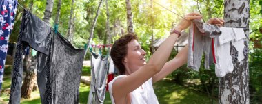 Açık havada güneşli bir günde ev bahçesinde broşlarla huş ağacının çamaşır ipine yeni yıkanmış aile giysileri asan güzel beyaz bir kadının samimi gerçek hayat portresi.