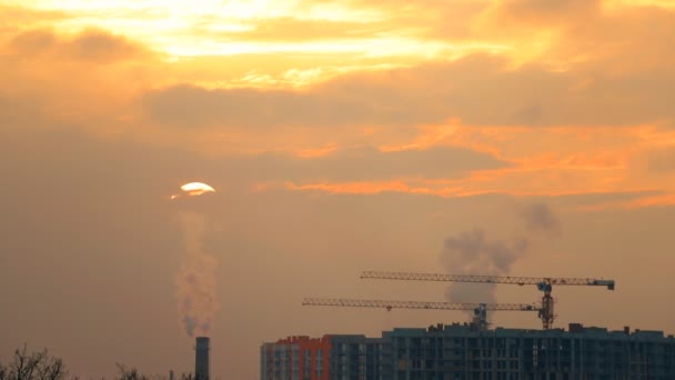 Vista al tramonto dello skyline dell'orizzonte della città con fumaioli tubi, gru in cantiere e sole nascosto nelle nuvole. Scena urbana serale. Inquinamento ambientale da CO2 — Video Stock