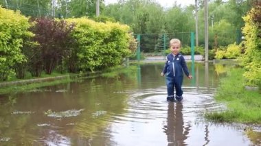 Küçük sevimli, oyuncu, beyaz, sarışın çocuk açık hava bahçesinde mavi su geçirmez pantolon ve lastik çizmelerle kirli su birikintisinde zıplayarak eğleniyor. Mutlu çocukluk kavramı