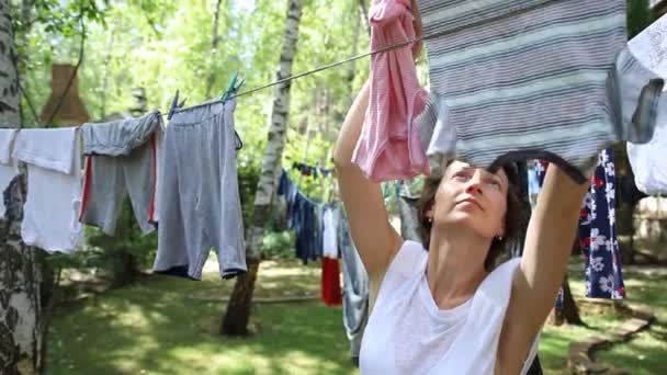 在阳光明媚的户外，年轻美丽迷人的高加索女人用别针在自家院子里的桦树晾衣绳上挂着新鲜的洗过的家居服 — 图库视频影像