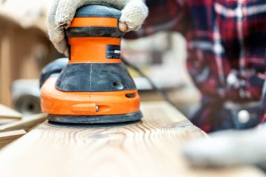 Closeup professional carpenter hand grinding raw wood plank with orbital sander electric machine in carpentry diy workshop. Detail of furniture restoration renewal. Power tool and equipment concept, clipart