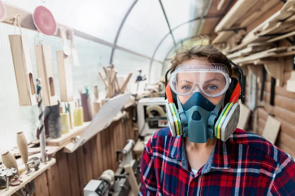 Young beautiful handy professional female carpenter close-up portrait wearing ppe respirator goggles and ear protection in carpentry diy workshop. Confident engineer. Women male hobby at workbench.