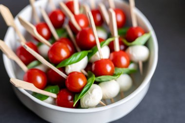 Beyaz kasede servis edilen kiraz domatesli, mozarella toplu ve fesleğenli taze kanepe şişleri. Renkli aperatifler İtalyan caprese tarzını yansıtıyor. Parti atıştırmalıkları ya da şenlik yemekleri için mükemmel..