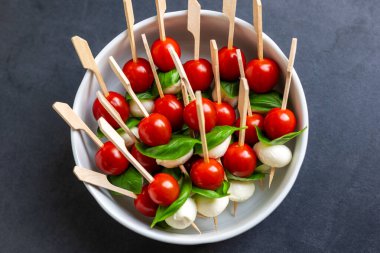 Beyaz kasede servis edilen kiraz domatesli, mozarella toplu ve fesleğenli taze kanepe şişleri. Renkli aperatifler İtalyan caprese tarzını yansıtıyor. Parti atıştırmalıkları ya da şenlik yemekleri için mükemmel..