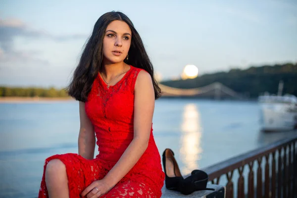 Una chica vestida de rojo está sentada en la cerca del terraplén del río. . — Foto de Stock